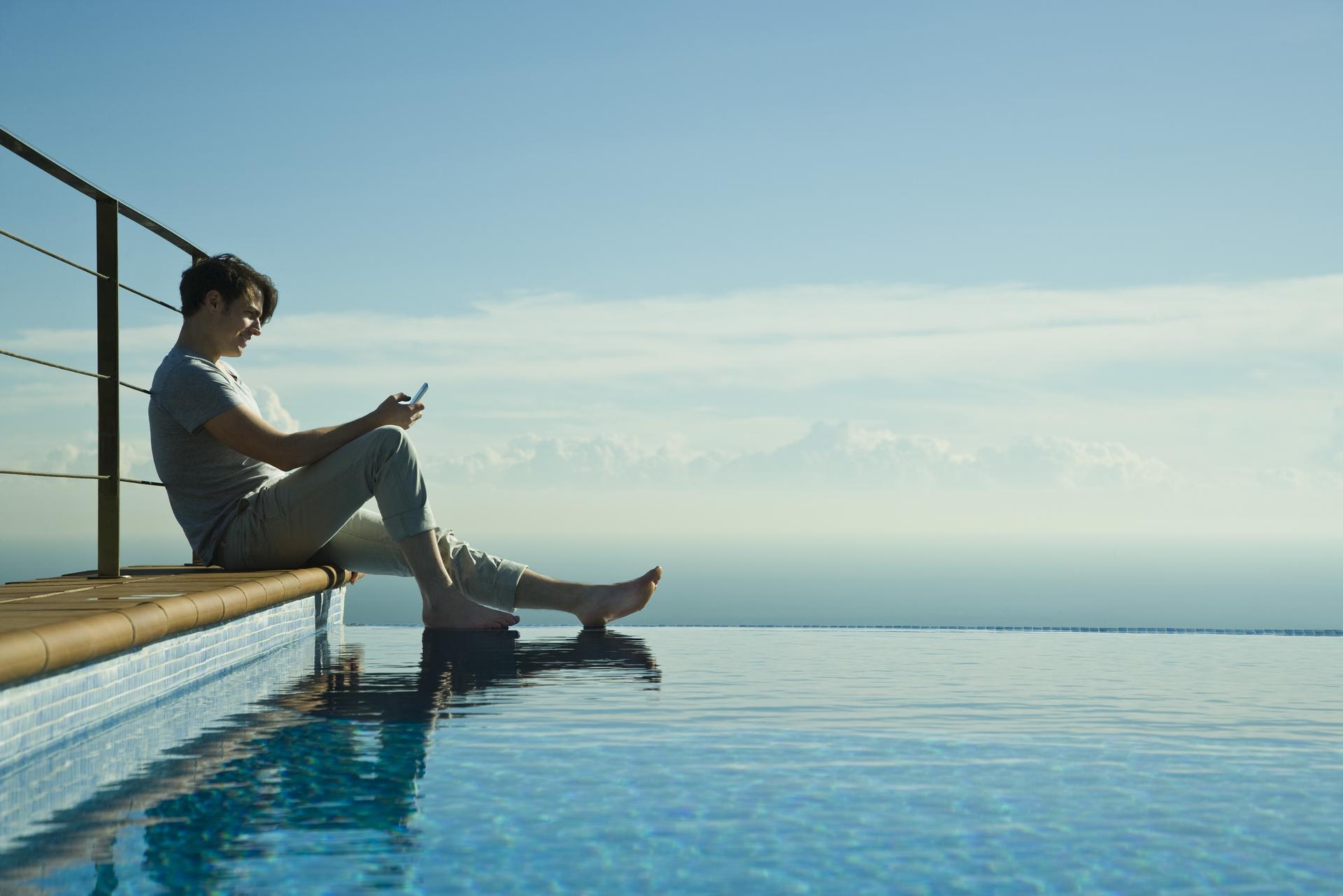 5486 Man sitting on edge of infinity pool, text messaging with cell phone