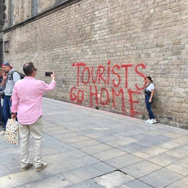Turistas fotografiándose frente a la pintada que dice %22turistas idos a casa%22 en Barcelona (Europa Press)